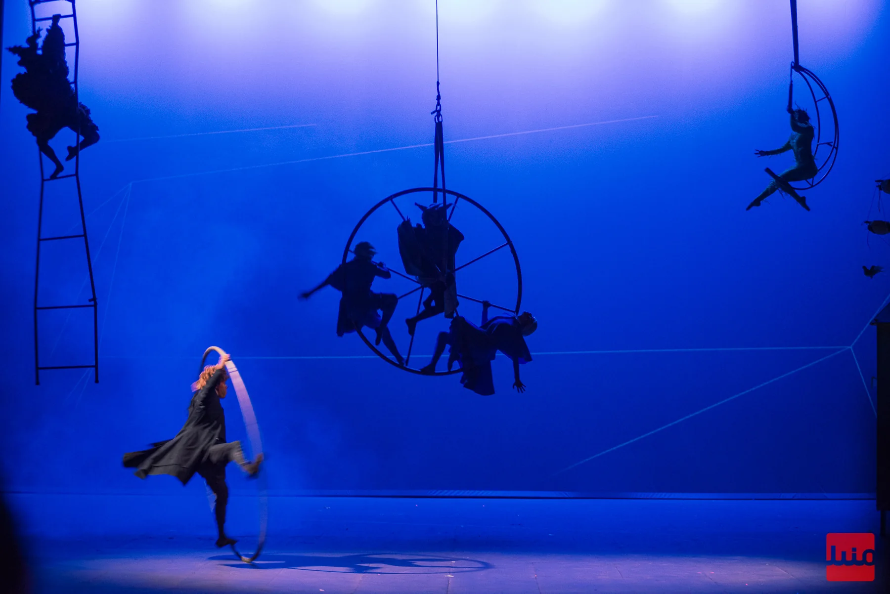 Intérpretes sobre aparatos aéreos y una bailarina ocupan distintos planos del escenario.
